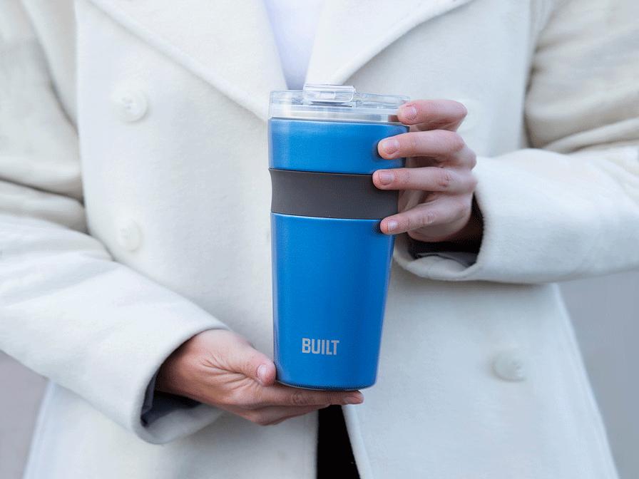 Person in a white coat holding a blue BUILT insulated tumbler with a clear lid.