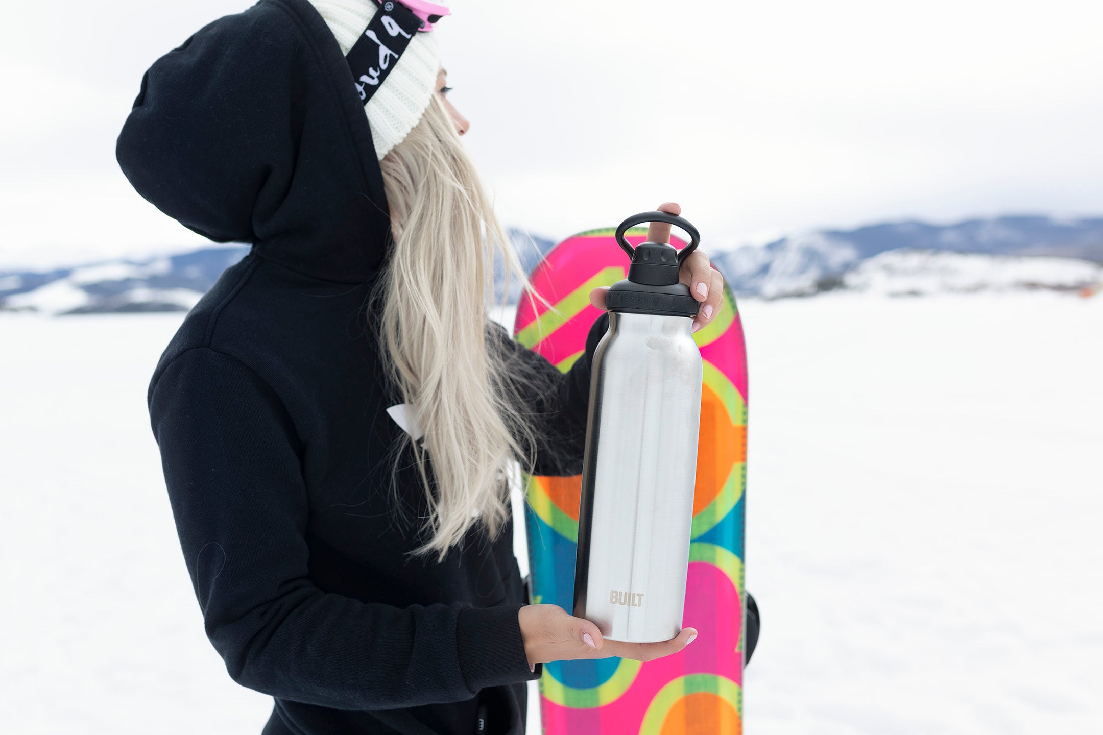 snow boarder holding a white water bottle tumbler in the snow looking at a mountain