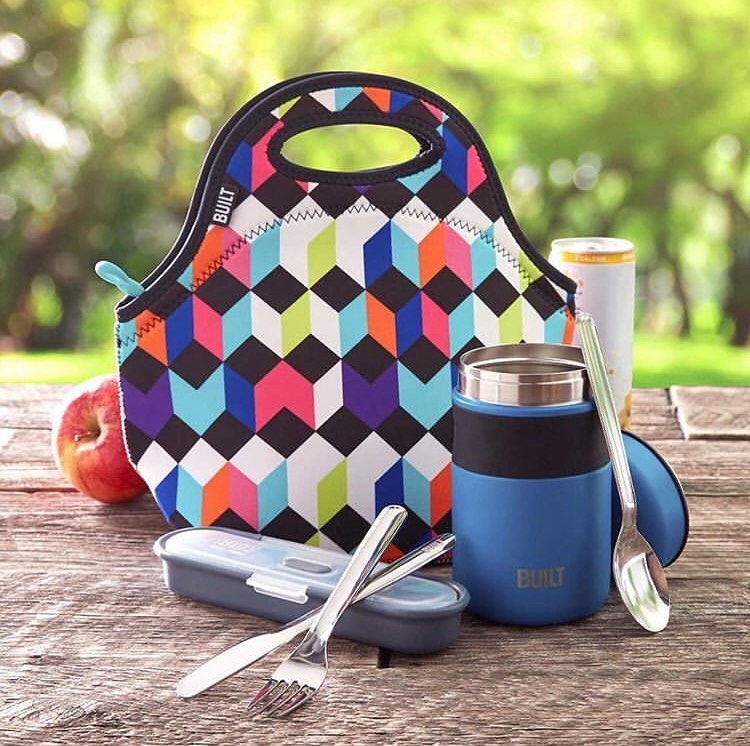 lunch bag with fork and knife and spoon utensils and food jar outdoors on a picnic table with apple and drink